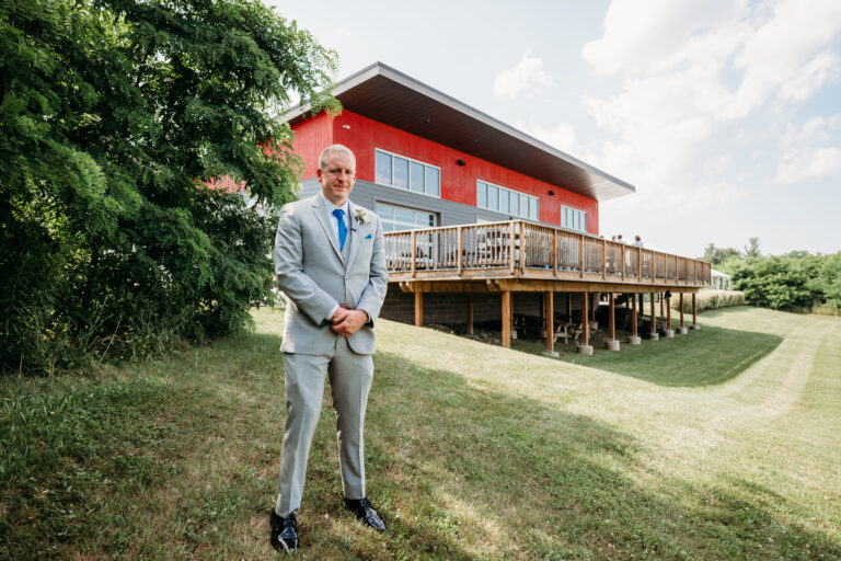 A man in a gray suit stands on a grassy lawn in front of a red building with a wooden deck.