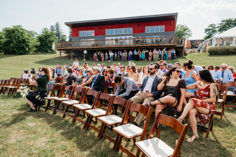 Outdoor ceremony with guests seated on wooden chairs on a grassy lawn in front of a red barn-style venue.
