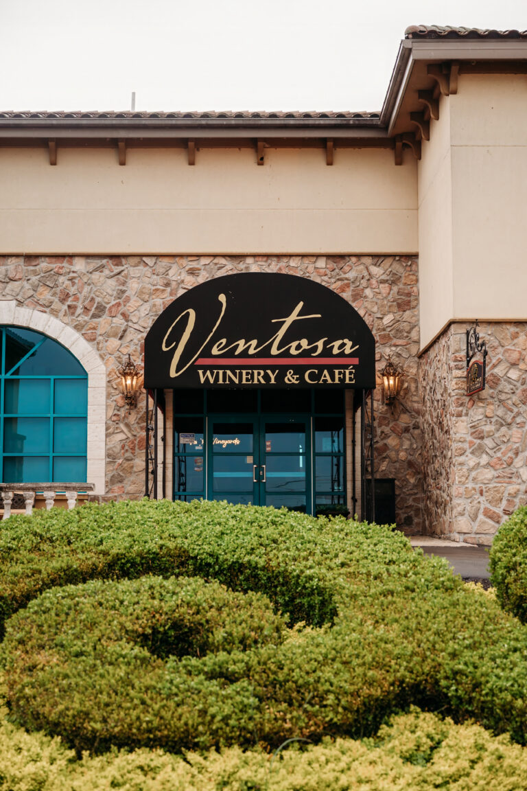 Exterior view of Ventosa Winery & Café with a stone facade, an arched turquoise window, and neatly trimmed hedges in the foreground.