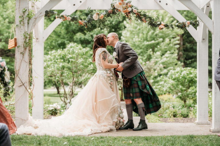 Bride in a pale pink wedding gown and groom in a tartan kilt kiss under a white garden arch.