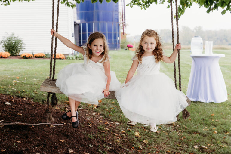 Two young girls in white dresses swing on a wooden swing outdoors, smiling at the camera.