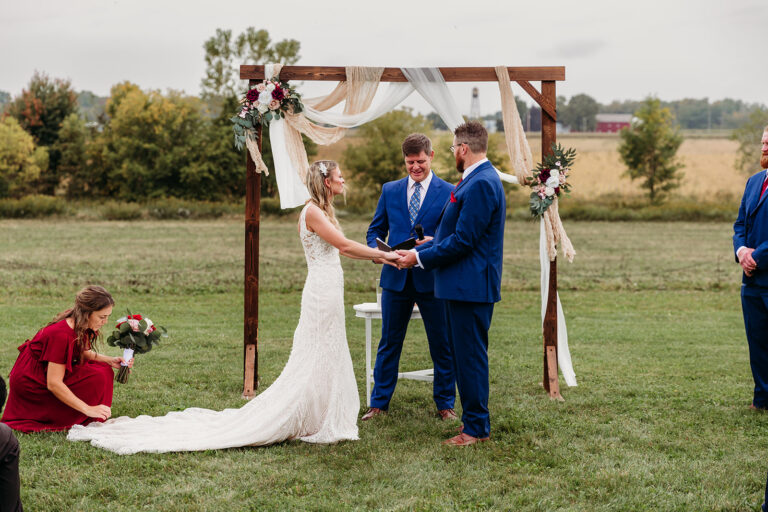 Bride and groom exchange rings under a flower-decorated wooden arch during an outdoor wedding ceremony.