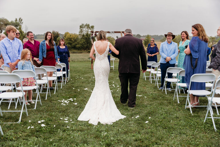 A bride in a white wedding gown walks down an outdoor grassy aisle with a man in a black suit as guests sit in white chairs on either side.