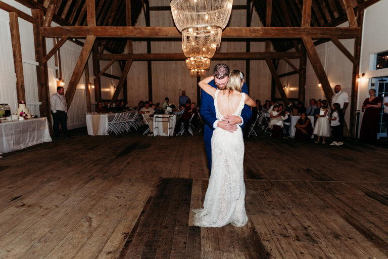 Bride and groom dancing in a rustic barn wedding reception under a chandelier, with guests seated at tables along the walls.