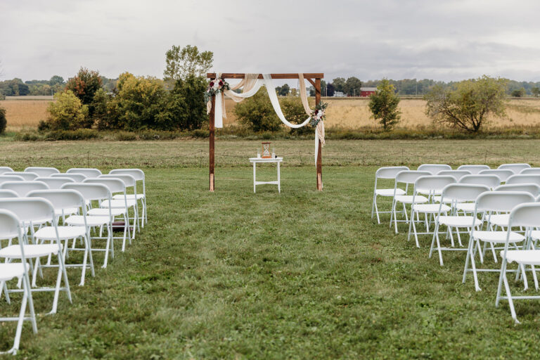 Outdoor wedding ceremony setup on a grassy field with rows of white folding chairs facing a wooden arch draped in white fabric and greenery.