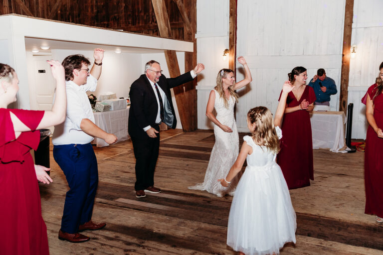Wedding guests in formal attire dance on a wooden barn floor, with a young girl in a white dress at the center.