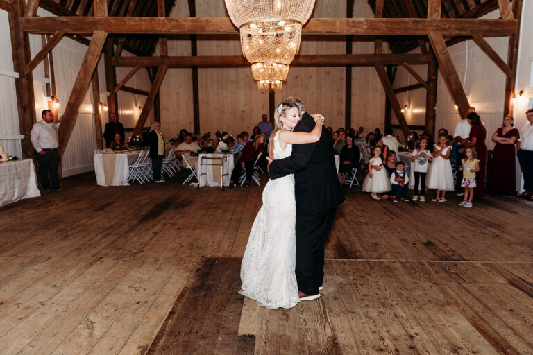 A bride and groom dance together in a rustic wooden venue under a chandelier, with guests seated at tables watching.