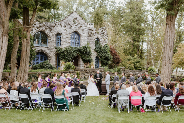 Outdoor wedding ceremony in front of a stone church with arched windows, as guests sit in rows watching the couple.