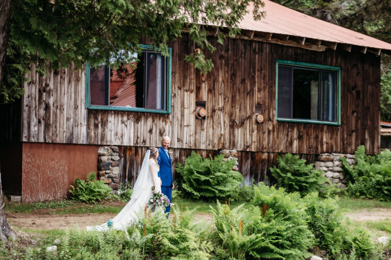 A bride in a white wedding dress and a groom in a blue suit walk along a path in front of a rustic wooden house surrounded by greenery.