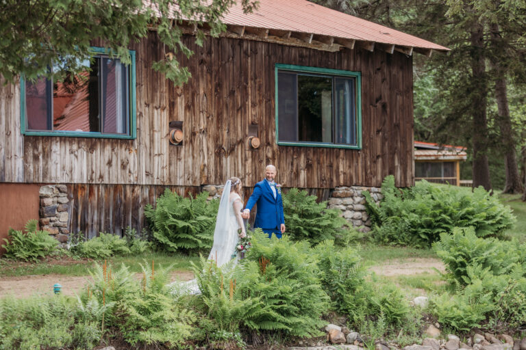 Bride and groom walk together in front of a rustic wooden cabin with green-trimmed windows, surrounded by greenery.