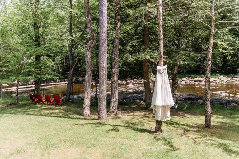 A white dress hangs from a tree in a sunlit grassy yard with a stream and red chairs in the background.