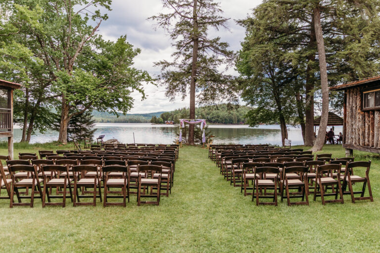 Rows of wooden chairs on a grassy lawn face a lakeside wedding arch with trees and water in the background.