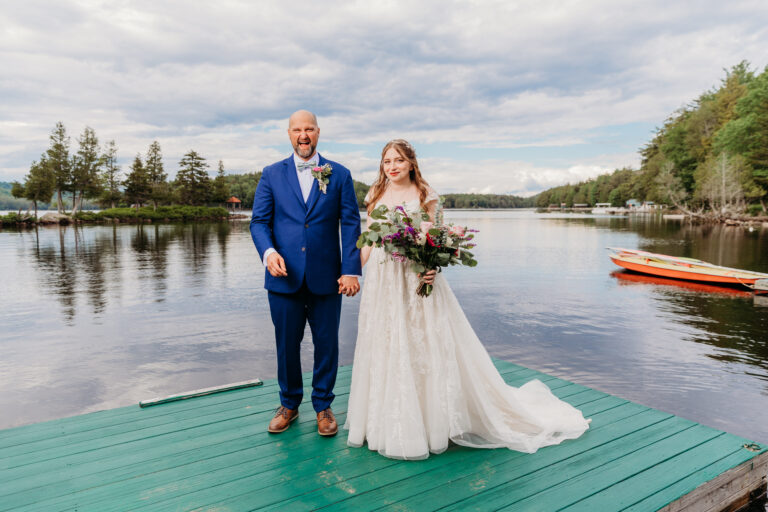 Bride in a white wedding gown and groom in a blue suit stand on a green dock by a calm lake with trees in the background.