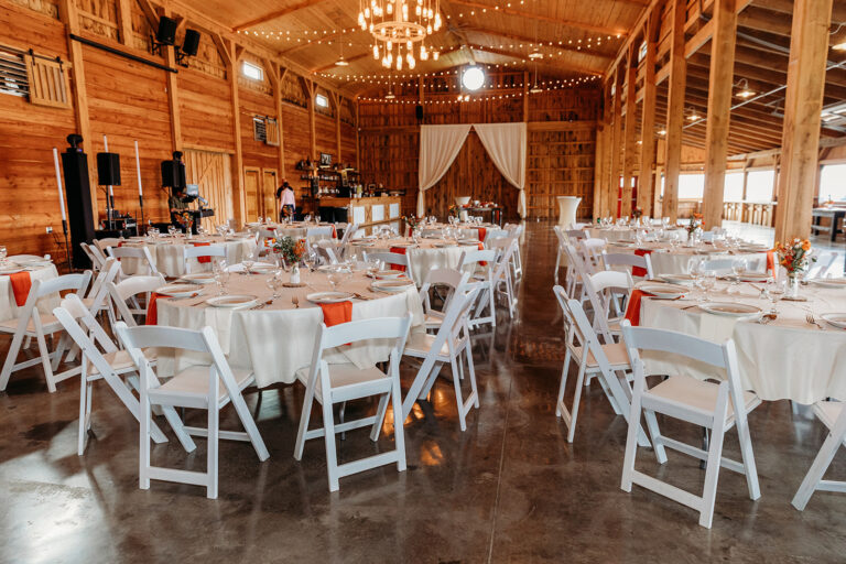 Rustic barn wedding reception setup with round tables covered in white linens, white folding chairs, and warm chandelier lighting.
