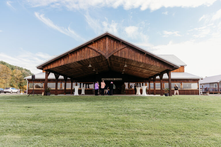 Open-air wooden pavilion with a large gabled roof sits on a green lawn, with people gathered inside under the shade.