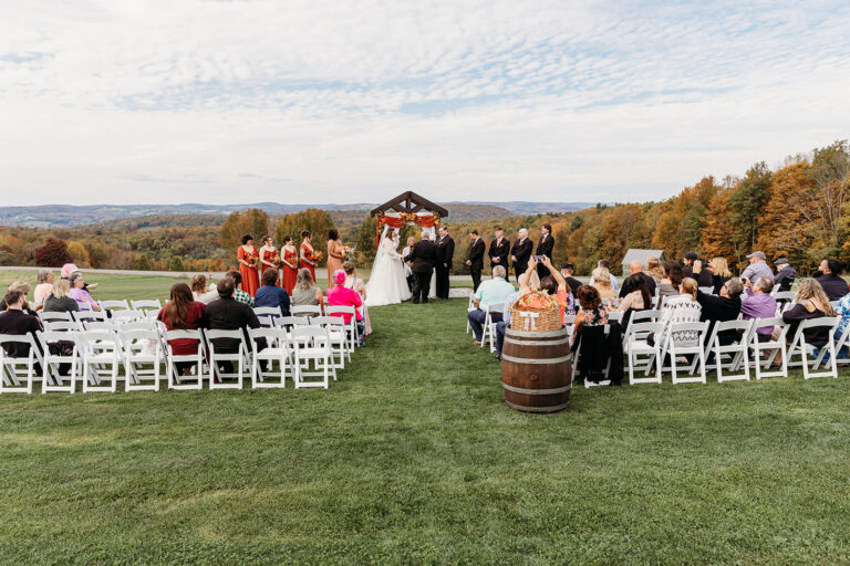 Outdoor wedding ceremony on a grassy field with white folding chairs facing a wooden arch, surrounded by autumn trees and guests.