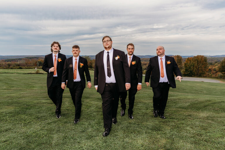 Five men in black suits with orange ties walk toward the camera across a grassy field under a cloudy sky.