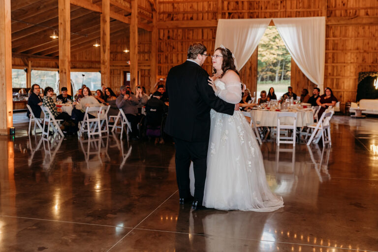 Bride and groom dance on a wooden floor at a rustic barn wedding reception, with guests seated at tables in the background.