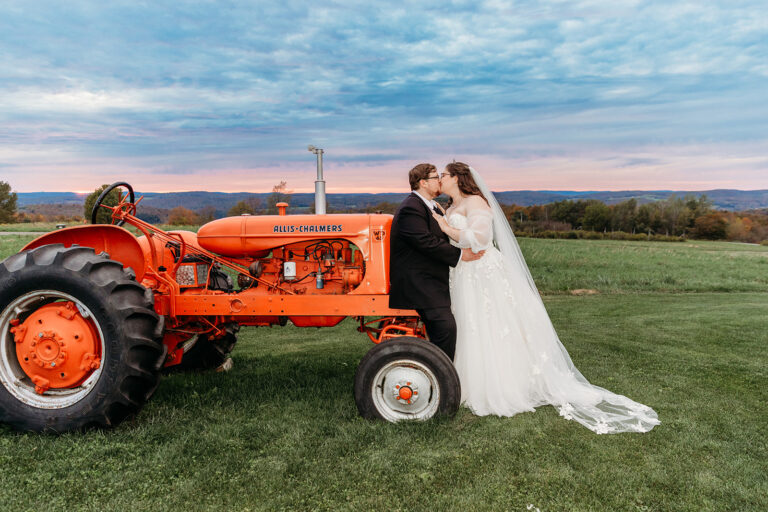 A bride in a white wedding dress and a groom kiss beside a vintage orange tractor in a green field.