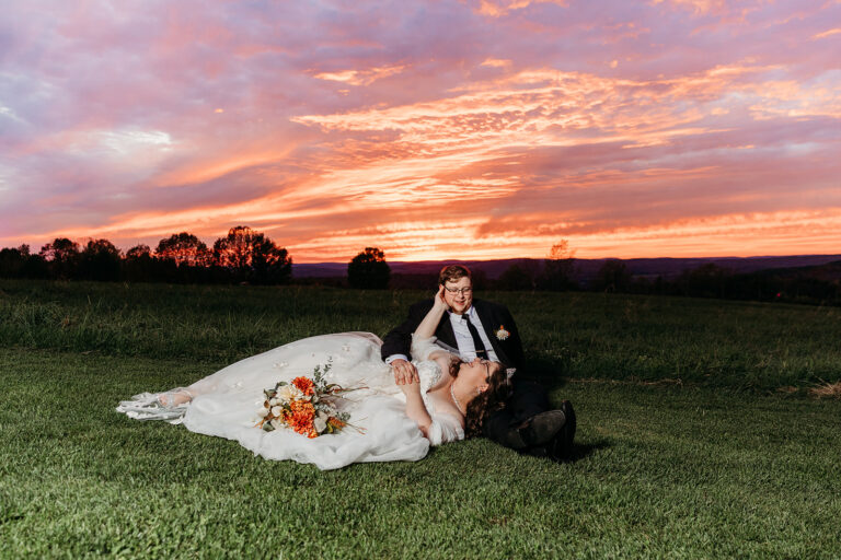 A bride in a white wedding dress lies on the grass beside her groom in a dark suit, with a vivid orange-pink sunset in the background.