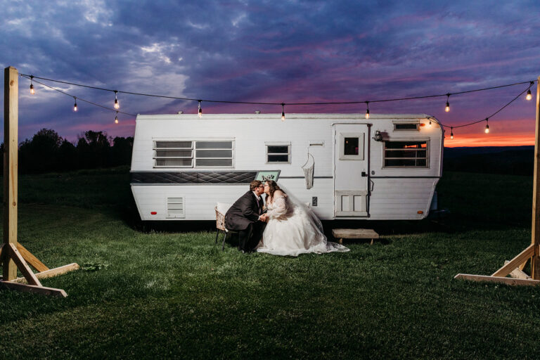 A bride and groom sit in front of a white camper trailer on a grassy field at sunset, with string lights overhead.