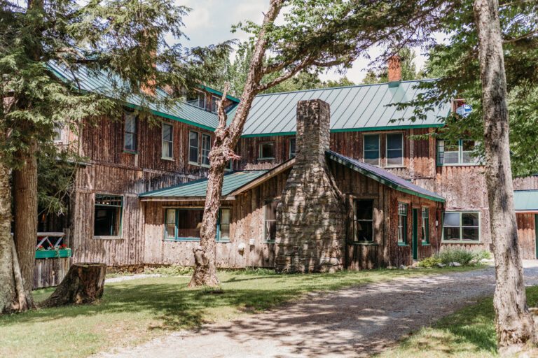A brick building with a green metal roof sits among tall trees, featuring a stone chimney and a wooden entrance along a dirt path.