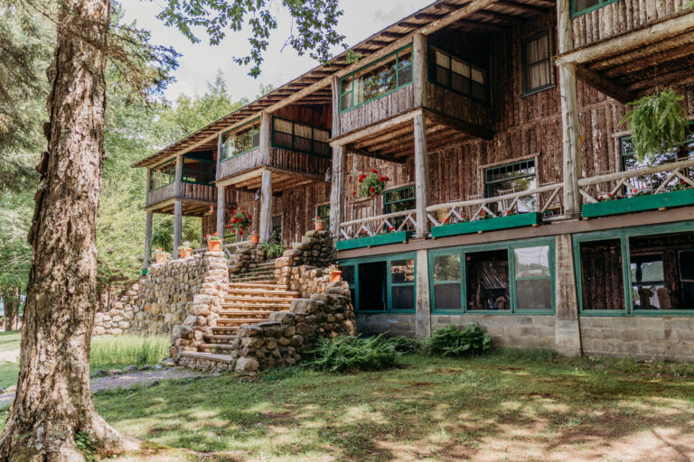 A rustic two-story wooden lodge with wraparound balconies, a stone staircase, and green trim sits on a grassy lawn among trees.