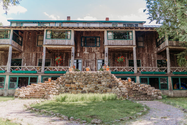 Rustic two-story log cabin with a central stone staircase leading to the front entrance and a wraparound porch, set among trees.
