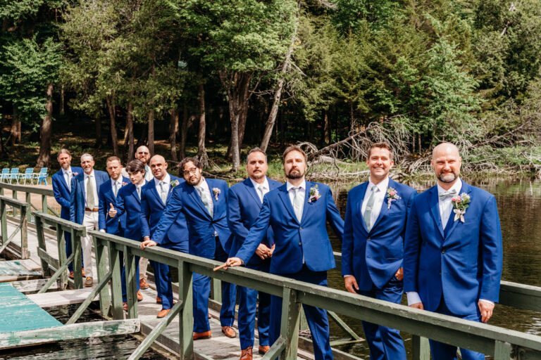 Groomsmen in matching blue suits stand on a wooden dock by a lake with a forest backdrop.