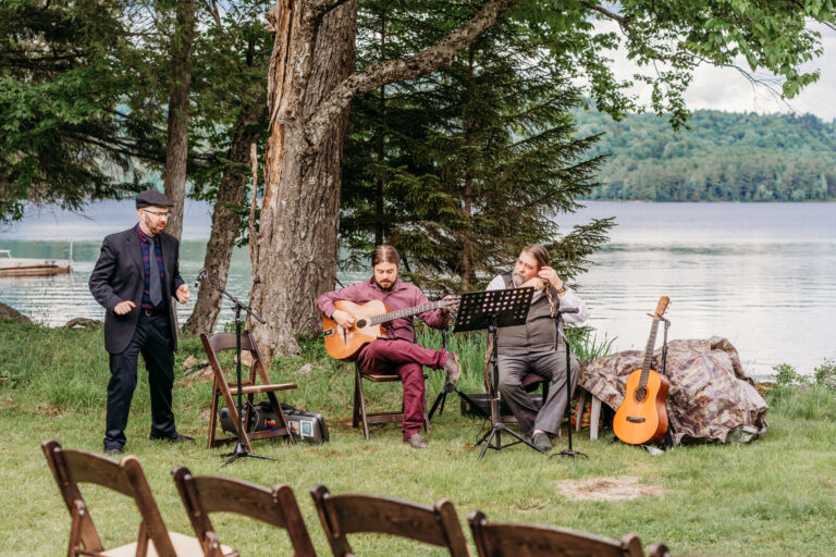 A small group of musicians playing guitars outdoors by a lakeside, with trees, logs, and empty wooden chairs in the foreground.