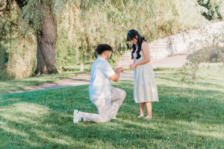 A man kneels on the grass proposing to a woman in a white dress in a park.