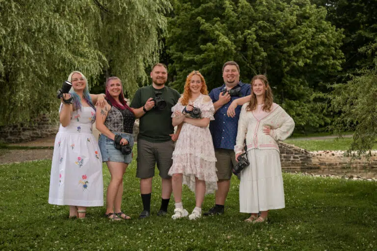 Six adults stand together on a grassy park lawn, posing and smiling for the camera.