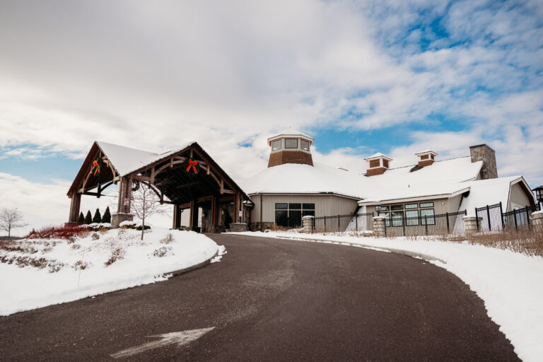Snow-covered lodge with a curved driveway leading to the entrance, featuring a central round tower and rustic stone-and-wood exterior under a partly cloudy sky.