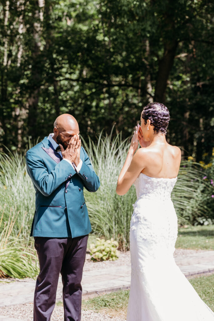 A bride in a strapless white wedding dress and a man in a teal blazer stand outdoors in a garden, both covering their mouths with their hands.