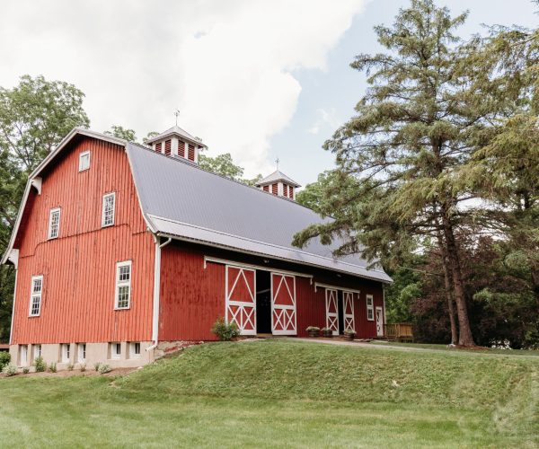 A rustic red barn with a gray roof surrounded by lush greenery and tall trees on a cloudy day.