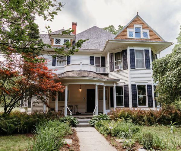 A Victorian-style house stands surrounded by lush greenery, featuring white siding, a wraparound porch, and dark shutters under a cloudy sky.
