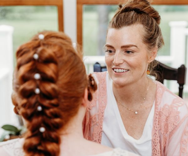 Two people indoors, one with braided hair facing away, another smiling, wearing a white shirt and pink lace garment, in conversation.