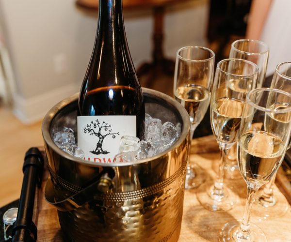 A wine bottle in an ice bucket is surrounded by six filled champagne flutes on a wooden surface. A person is partially visible.