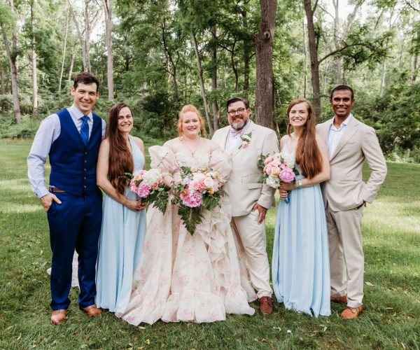 A group of six people dressed for a formal outdoor event, standing on grass with a wooded background, holding bouquets.