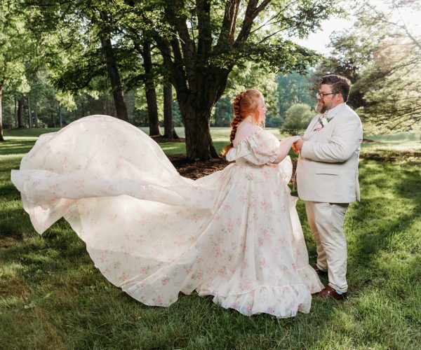 A person in a floral gown holds hands with another person in a suit, standing amidst lush green trees in a sunlit park.