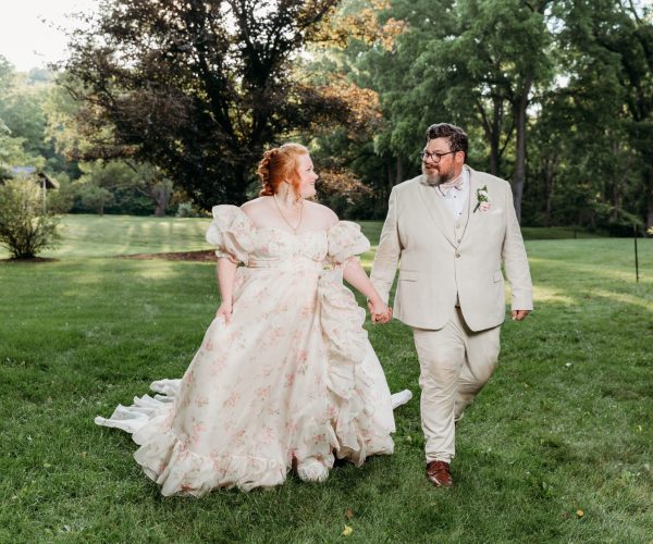 A couple in formal attire walks hand in hand on lush green grass, surrounded by leafy trees and a serene garden setting.
