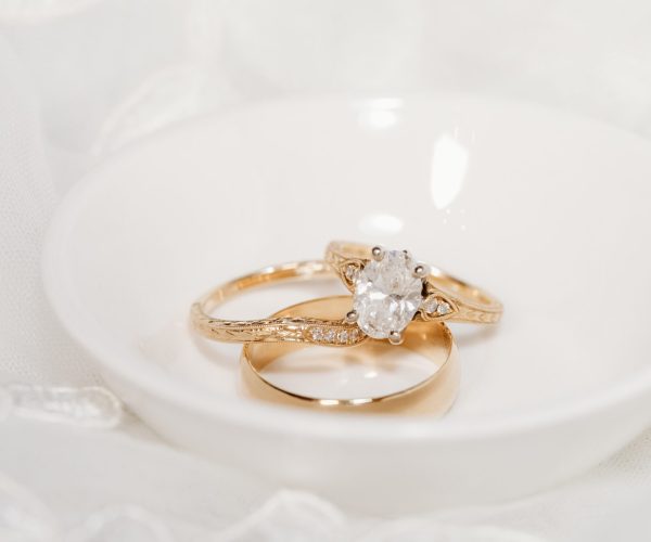 Two gold rings, one with a diamond, rest elegantly in a white dish against a soft fabric backdrop, symbolizing commitment and elegance.