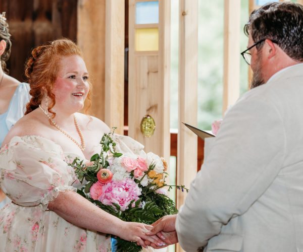 A person in a floral dress smiles, holding hands with another person in a formal setting, accompanied by a person holding flowers.