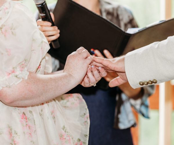 A couple holds hands during a wedding ceremony, with a person officiating and holding a microphone and a black folder.