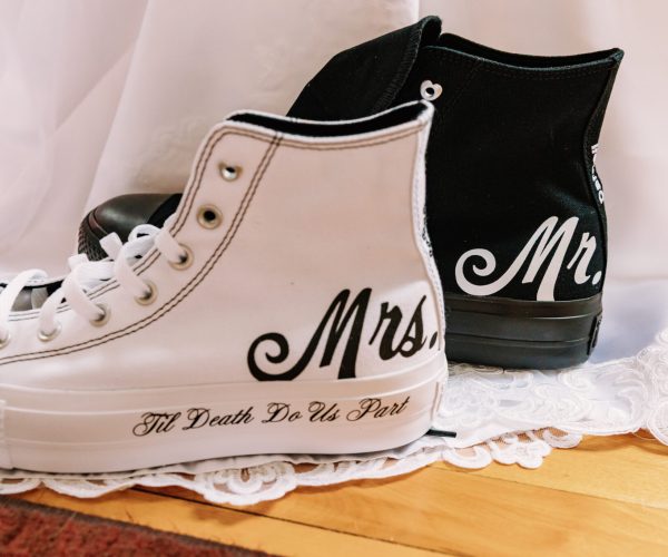 A pair of personalized black and white sneakers labeled "Mr." and "Mrs." rests on delicate lace, featuring wedding-themed text.
