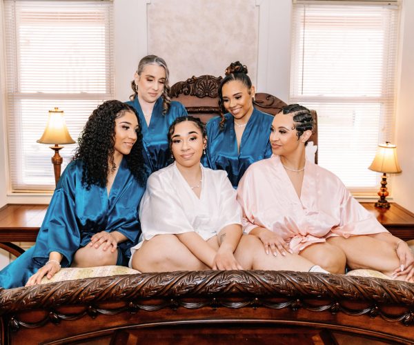 Five persons in colorful robes sit on a bed. The room features classic wooden furniture and soft lighting from table lamps.