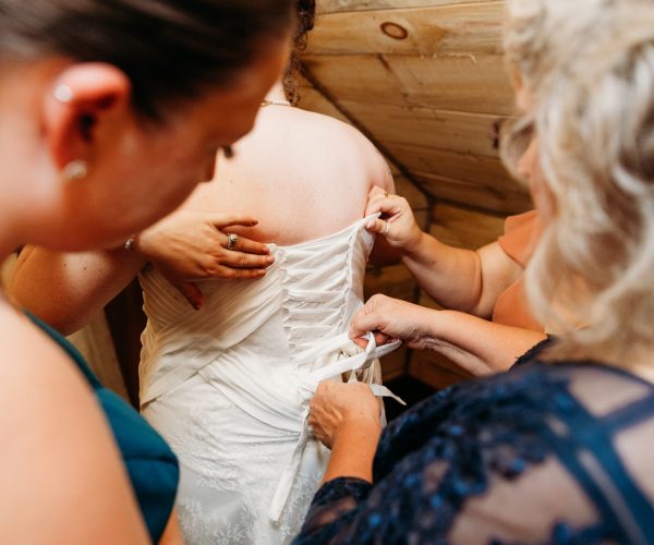Three people help adjust the back of a wedding dress in a warmly lit wooden room, focusing on the intricate lacing details.