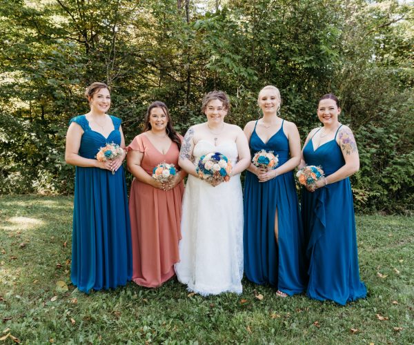 Five people in formal dresses stand in a grassy area holding flowers, surrounded by lush green trees in the background.