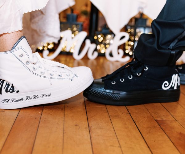 Two people wearing custom sneakers labeled "Mrs." and "Mr." face each other. Background features illuminated "Mr & Mrs" sign on wooden floor.