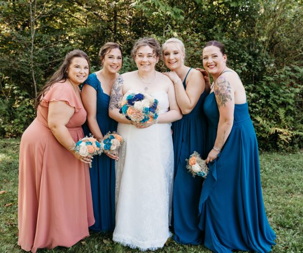 Five people in formal dresses stand smiling, holding bouquets, in a lush outdoor setting with trees in the background.